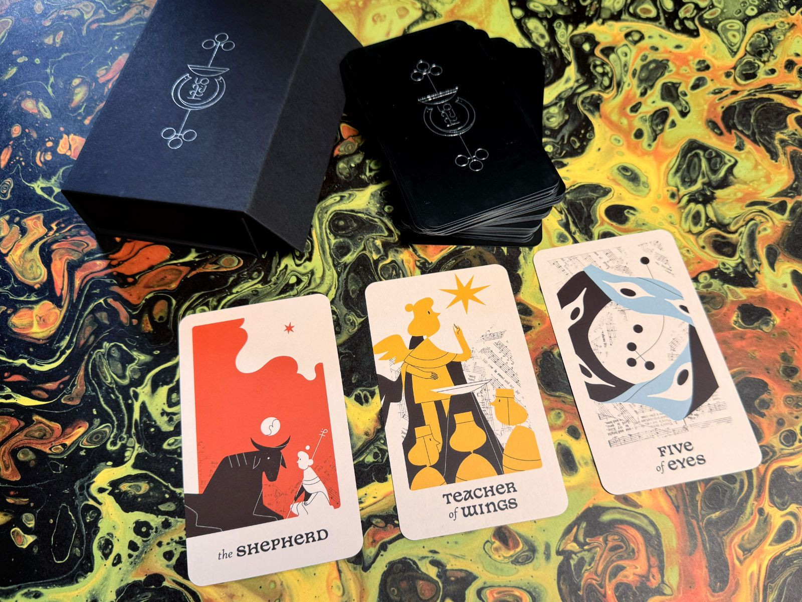 A lovely promotional shot from Knave of Cups. You can see the box and back of the cards with foil stamping, and three cards are laid out in a spread in the front — The Shepherd, the Teacher of Wings, and the Five of Eyes.